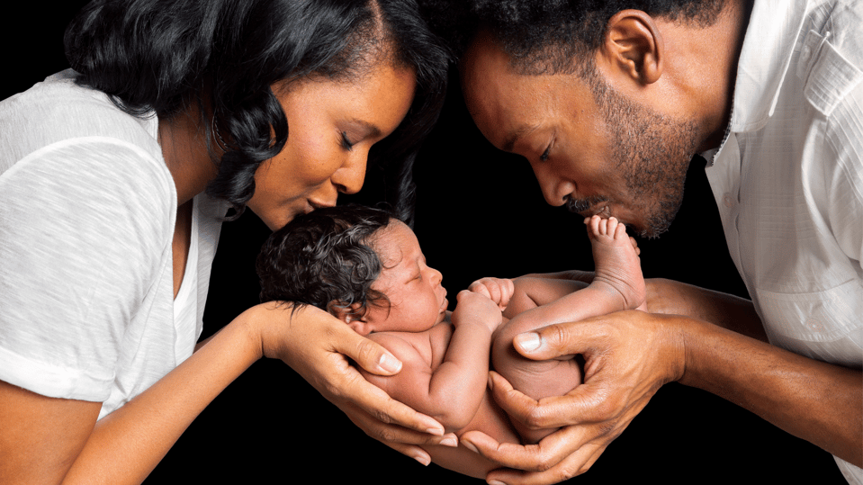 Two parents holding a newborn child in both of their hands and kissing the baby’s forehead.
