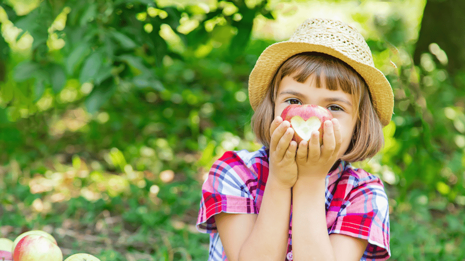 A child in a straw hat holding an apple with a bite in the shape of a heart. 